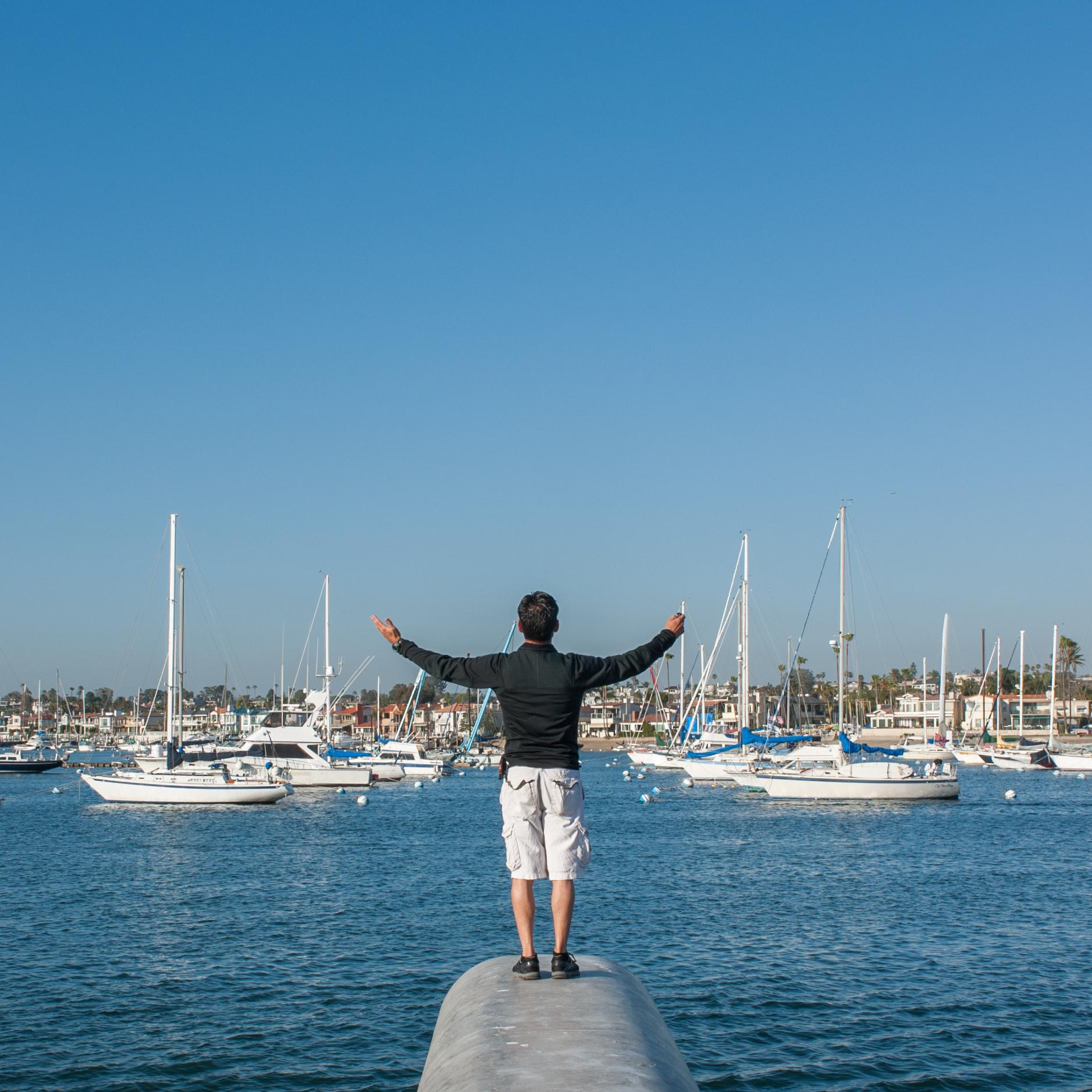 Man standing with arms outstretched on in front of a harbor with boats docked on a sunny day